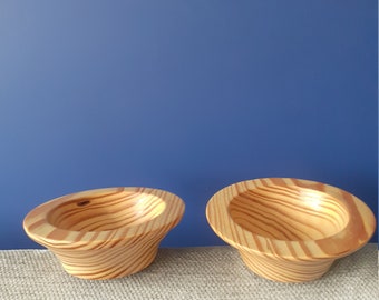May include: Two wooden bowls with a natural wood grain pattern. The bowls are light brown with darker brown stripes. The bowls are sitting on a light brown textured surface.