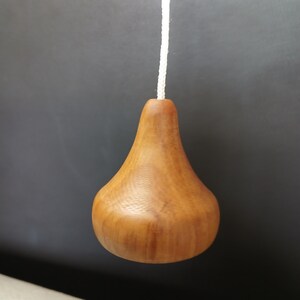 Puede incluir: Un tirador de luz de madera en forma de pera con un acabado de veta de madera natural de color marr&oacute;n claro, unido a un cord&oacute;n trenzado blanco fino. El objeto presenta una base redondeada que se estrecha hacia un cuello donde va el cord&oacute;n.
