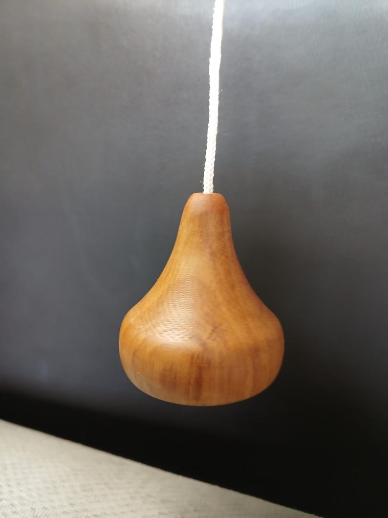 Puede incluir: Un tirador de luz de madera en forma de pera con un acabado de veta de madera natural de color marr&oacute;n claro, unido a un cord&oacute;n trenzado blanco fino. El objeto presenta una base redondeada que se estrecha hacia un cuello donde va el cord&oacute;n.