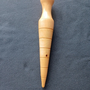 May include: A light-coloured wooden massage tool with a rounded top and a tapered, pointed end. The tool has a series of horizontal grooves and a small, dark spot. The background is a solid blue fabric.