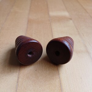 Peut inclure: Deux boutons en bois brun fonc&eacute; de forme cylindrique avec un trou au centre. Les boutons pr&eacute;sentent une s&eacute;rie de rainures horizontales sur leur circonf&eacute;rence. Le fond est une surface en bois brun clair.