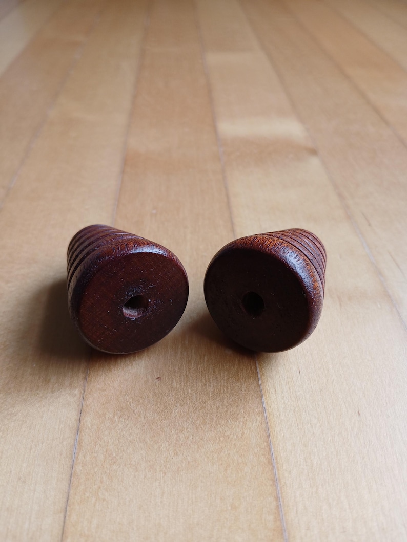 Peut inclure: Deux boutons en bois brun fonc&eacute; de forme cylindrique avec un trou au centre. Les boutons pr&eacute;sentent une s&eacute;rie de rainures horizontales sur leur circonf&eacute;rence. Le fond est une surface en bois brun clair.
