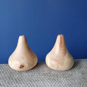 Pode incluir: Dois pequenos vasos de madeira em forma de gota com um acabamento liso e claro. Cada vaso apresenta um pescoço estreito e uma base larga e arredondada, exibindo padrões naturais de veios de madeira e um pequeno nó na peça da esquerda.