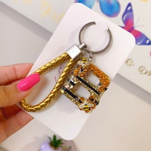 May include: Gold keychain with a braided rope strap and a letter B charm. The charm is decorated with glitter, black stripes, and small bee accents.