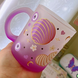 May include: A frosted purple mug with a handle. The mug features a design of seashells, stars, hearts, and evil eyes. The evil eyes are blue and white.
