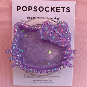Puede incluir: Un soporte para teléfono PopSockets morado brillante con forma de cara de gato de dibujos animados con un lazo. El PopSocket está en una tarjeta blanca con el nombre de la marca y el texto "Non Textured Surfaces Only".