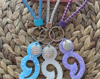 May include: Three resin keychains in blue, white, and purple, each with a braided strap and a silver keyring. The keychains are shaped like a figure eight with glitter and the text "She believed she could so she did."
