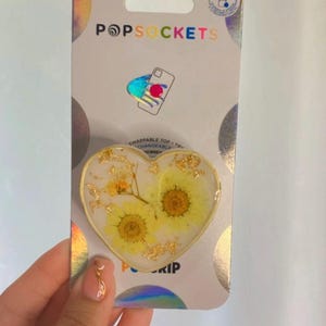 May include: Heart-shaped PopSockets phone grip featuring yellow flowers and gold flakes encased in clear resin. The grip is mounted on a holographic backing with the PopSockets logo.