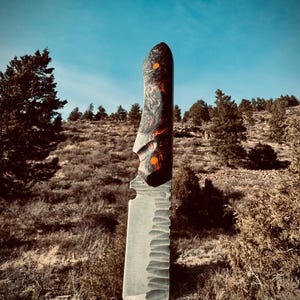 May include: A handmade knife with a black and red marbled handle and a silver blade with a serrated edge. The knife is resting on a piece of wood.