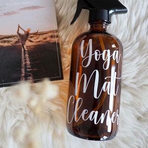 Könnte beinhalten: Eine braune Glas-Sprühflasche mit schwarzer Sprühdüse. Die Flasche hat ein weißes Etikett mit der Aufschrift "Yoga Mat Cleaner".