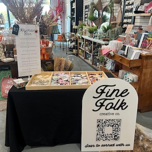 May include: A shop display with a "Keychain Bar" sign and instructions. A table showcases keychains and accessories. A sign for "Fine Folk creative co." with a QR code is in the foreground.
