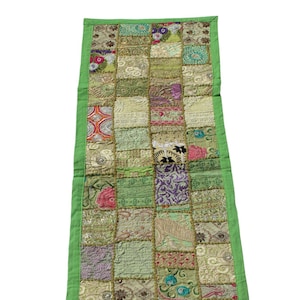 May include: A green patchwork table runner with a variety of colorful embroidered designs. The runner is made of different fabrics and has a green border.