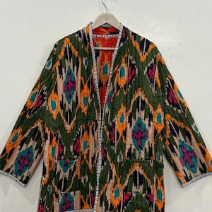 Può includere: Giacca trapuntata con apertura frontale e un vivace motivo geometrico nei toni del verde, arancione, rosa e turchese. La giacca ha una fodera rossa ed è rifinita con un bordo a righe bianche e nere. Appesa a un muro bianco.