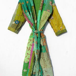 May include: A patchwork robe in shades of green, pink, and gold, with a tie belt. The robe is made of quilted fabric with various patterns and textures. The robe is hanging on a wooden hanger.