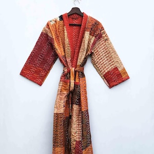 May include: A long, patchwork robe in shades of red, orange, and brown hangs against a white background. The robe features a tie belt and is made of various fabric squares stitched together. The sleeves are wide and the robe is hanging on a wooden hanger.