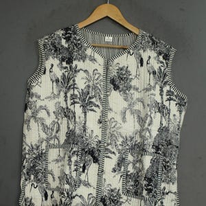 Könnte beinhalten: Eine ärmellose, gesteppte Weste mit einem schwarz-weißen Toile-Druck von tropischen Palmen, Zebras und Kranichen. Das Kleidungsstück hat einen V-Ausschnitt, eine Knopfleiste vorne, zwei aufgesetzte Taschen und schwarz-weiß gestreifte Kanten.