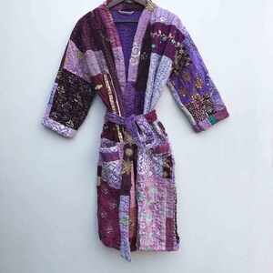 May include: A patchwork robe in shades of purple, lavender, and burgundy, with a matching belt. The robe features a quilted design with various fabric patterns and textures, including embroidered details. It has pockets and is hanging on a wooden hanger.