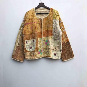 May include: A patchwork jacket in shades of yellow, brown, and cream, with a quilted texture. The jacket features two front pockets and floral embroidery. It is hanging on a wooden hanger against a white background.
