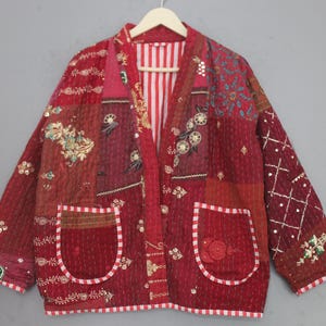 May include: A red patchwork jacket with embroidered floral and geometric designs. The jacket has two front pockets and a striped lining. The sleeves and body are decorated with detailed stitching and embellishments. The jacket is hung on a wooden hanger.