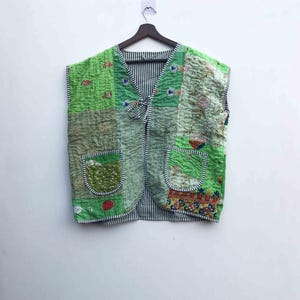 May include: A patchwork vest in shades of green, featuring various textured fabrics and patterns. The vest has two front pockets and a striped lining. The front closure is a tie. The vest is sleeveless and hangs on a wooden hanger.