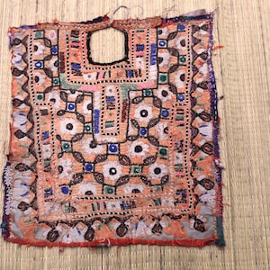 May include: A square, hand-stitched textile with an open neckline. The fabric is primarily orange with intricate patterns in white, black, blue, and green. The edges are trimmed with a purple and red border. The textile appears to be vintage or handmade.