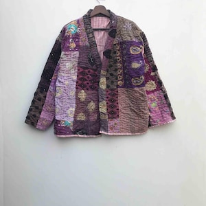 Op de afbeelding: Een patchworkjas in tinten paars, bruin en lavendel, met ingewikkelde borduursels en paisley patronen. De gewatteerde jas heeft een gestreepte voering, wat een unieke en bohemien uitstraling creëert.