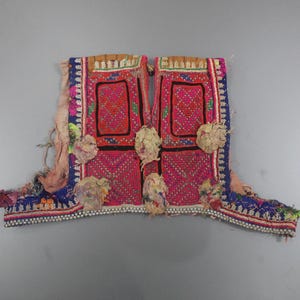 Può includere: Un tessuto ricamato vibrante con un design centrale rosa e rosso. Il capo presenta finiture di perline blu e bianche, con accenti color crema ed elementi floreali decorativi. Il tessuto è probabilmente un gilet o un capo simile.