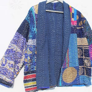 Op de afbeelding: Een patchwork jas in tinten blauw, paars en goud. De jas heeft een gewatteerd ontwerp met verschillende stofpatronen en texturen. De open voorkant en lange mouwen creëren een relaxte silhouet. De jas hangt aan een houten kleerhanger.