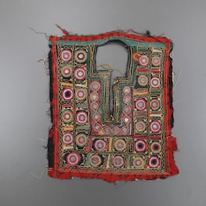 May include: A colorful, embroidered textile fragment with a central opening. The piece features a red border, black backing, and a pattern of squares and circles in various colors, including pink, yellow, and green. The design is symmetrical and intricate.