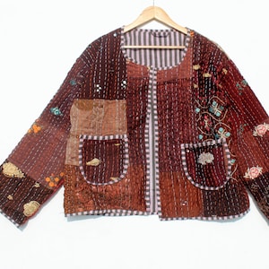 May include: A patchwork jacket in shades of brown, burgundy, and tan, with intricate stitching and embroidered details. The jacket features a round neckline, two front pockets, and a striped lining. The jacket is hanging on a wooden hanger.