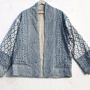 Könnte beinhalten: Blau-weiß gemusterte Jacke mit offenem Vorderteil. Die Jacke hat geometrische Designs, darunter Rauten, Streifen und florale Motive. Die Jacke hängt an einem Holzbügel vor weißem Hintergrund.