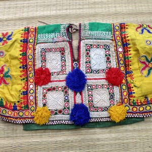 May include: A yellow and green textile garment featuring intricate embroidery, small circular mirrors, and vibrant red, blue, and yellow pom-poms. The piece displays geometric patterns, colorful thread work, and a central vertical opening.