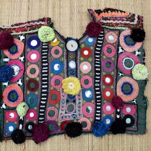May include: A handcrafted, dark green textile with colorful embroidery and embellishments. The design features multiple circular patterns in pink, blue, and orange, with small mirrors and pom-poms. The textile is likely a decorative piece or part of a garment.