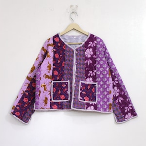 May include: A quilted jacket featuring a patchwork design in shades of purple, lavender, and white. The garment displays various patterns including floral motifs and tiger illustrations, with a V-neckline, long sleeves, and two front pockets.
