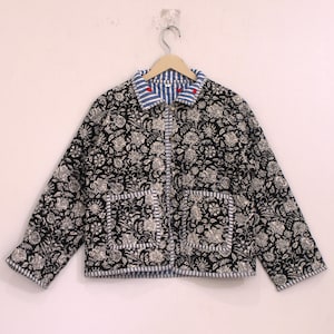 May include: A long-sleeved quilted jacket featuring a black and white floral block print pattern. The garment includes a blue and white striped collar, front placket, cuffs, and two square front pockets, accented by small red embroidered details.