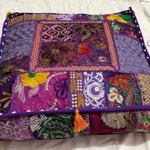 Könnte beinhalten: Ein quadratisches Bodenkissen mit Patchwork-Muster. Das Kissen ist aus violettem, rosa und gelbem Stoff mit aufwendigen Stickereien gefertigt. Das Kissen hat an den Ecken Quasten.