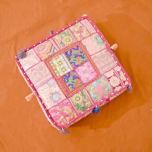 May include: A pink and white patchwork floor cushion with a square design and tassels. The cushion is made of different fabrics and has a variety of patterns.