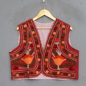 Könnte beinhalten: Eine rote ärmellose Weste mit aufwendiger Blumenstickerei in Orange-, Dunkelrot- und Hellgrüntönen. Das Kleidungsstück hat einen V-Ausschnitt und ist mit einem weißen Stoff mit dünnen vertikalen roten Streifen gefüttert, präsentiert auf einem Holzkleiderbügel.