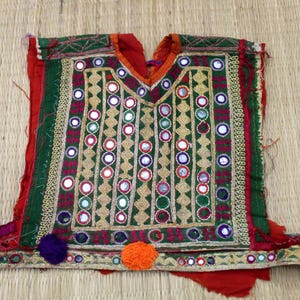 Puede incluir: Textil bordado de colores vivos, con diseño de cuello en V. La prenda presenta tela verde, roja y dorada con intrincados patrones y pequeños espejos redondos. Pompones decorativos en morado y naranja están adheridos al borde inferior.