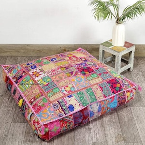 Könnte beinhalten: Ein großes, quadratisches Bodenkissen mit Patchwork-Design. Das Kissen weist eine lebendige Mischung aus Farben auf, darunter Rosa, Rot, Grün und Gold. Es hat einen rosa Rand und dekorative Quasten an den Ecken. Das Kissen liegt auf einem Holzboden.