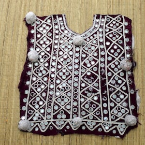 May include: A maroon textile with intricate white embroidery and small, round mirrors. The design features geometric patterns and is accented with small, fluffy, gray pom-poms. The garment is likely a vest or top, possibly of ethnic origin.