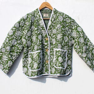 Puede incluir: Chaqueta acolchada con estampado floral verde y blanco, con cuello en V, dos bolsillos delanteros y botones de madera. La chaqueta está colgada de una percha de madera sobre un fondo blanco. El ribete de la chaqueta es blanco.