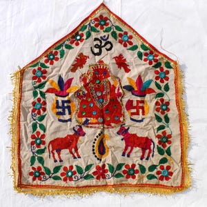 May include: Embroidered religious wall hanging with a cream background, featuring red and green floral patterns, a central deity figure, and the Om symbol. The design includes red cows, vases, and a gold fringe.