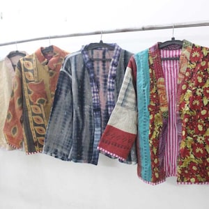 Może przedstawiać: Kolekcja kolorowych, patchworkowych kurtek kimono wiszących na metalowym wieszaku. Kurtki mają różne wzory, w tym kwiatowe, w paski i tie-dye. Paleta kolorów obejmuje odcienie niebieskiego, czerwieni, żółci i neutralne.