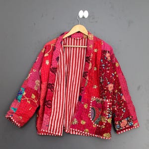Pode incluir: Casaco patchwork em tons de vermelho e rosa, com padrões florais e listrados. O casaco tem um corte folgado, um bolso e está pendurado em um cabide de madeira. O forro apresenta listras verticais vermelhas e brancas.