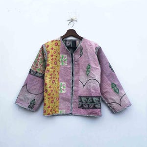 May include: A patchwork jacket in shades of lavender, yellow, and green, hanging on a wooden hanger. The jacket features a variety of floral and geometric patterns, with a black zipper and white trim. The sleeves are long and the jacket is open at the front.
