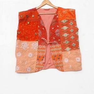 May include: A patchwork vest in shades of orange and peach, with a striped interior and a tie closure. The vest is made of various patterned fabrics, including floral and mirrored accents, and hangs on a wooden hanger.