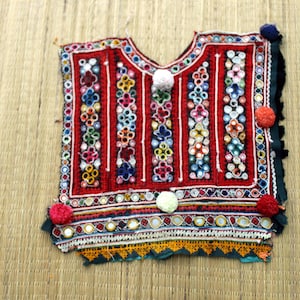May include: A vibrant, hand-stitched textile top with a red base, adorned with colorful floral patterns, mirrors, and pom-poms. The intricate design features vertical white stripes and a scalloped edge, showcasing a rich cultural heritage.