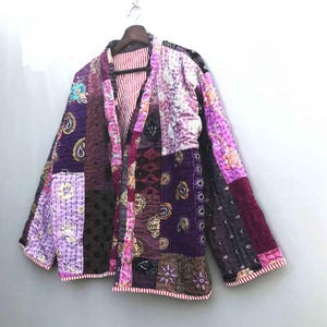 Può includere: Una giacca patchwork nei toni del viola, lavanda e bordeaux, con fodera a righe rosse e bianche. La giacca presenta una varietà di tessuti fantasia, tra cui disegni paisley e floreali. È appesa a una gruccia di legno.