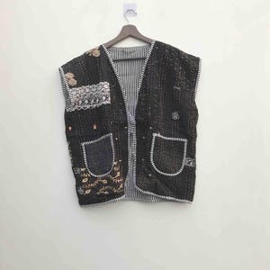 Peut inclure: Un gilet matelassé noir avec une bordure blanche, deux poches avant et un intérieur rayé. Le gilet présente des écussons brodés avec des motifs floraux et géométriques. Il est suspendu à un cintre en bois contre un mur blanc.